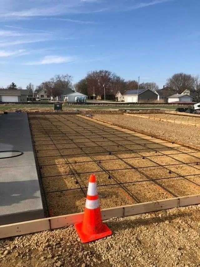 Construction site with a concrete slab, rebar grid, orange cone, and gravel. Buildings in the distance.