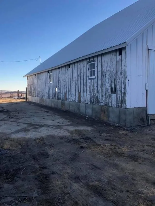 White barn with weathered paint and metal roof on a dirt lot under a clear blue sky.