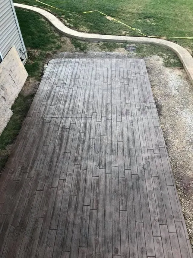 Stamped concrete patio with a weathered wood pattern. Surrounding area is gravel, grass, and a concrete walkway.