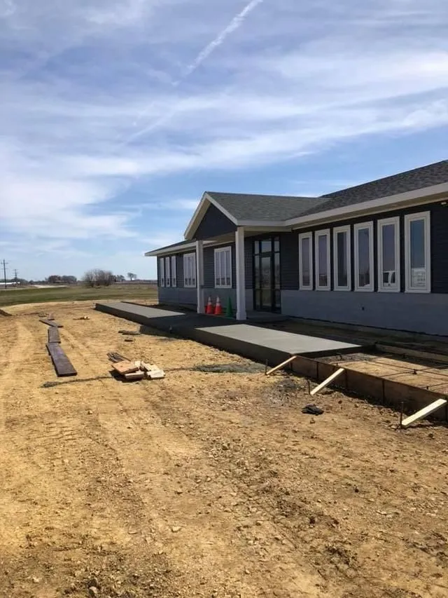 Building exterior under construction; gray siding, concrete patio, dirt, clear sky.