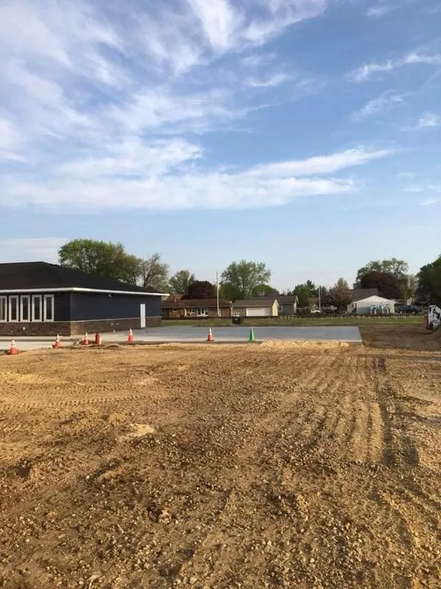 Construction site with newly poured concrete slab; blue building, cloudy sky.