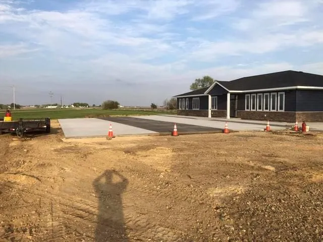 Construction site with asphalt and concrete path leading to a blue building; orange cones present.